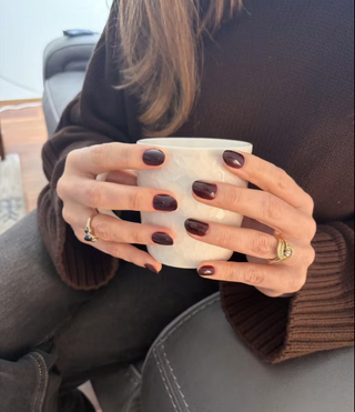 Person holding a white mug with dark nail polish and a brown sweater.