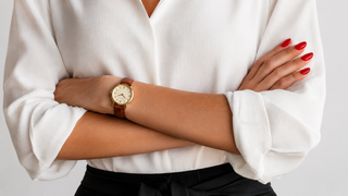 A close-up of a person wearing a gold-tone wristwatch with a brown leather strap, styled with a white blouse and black skirt against a light gray background.